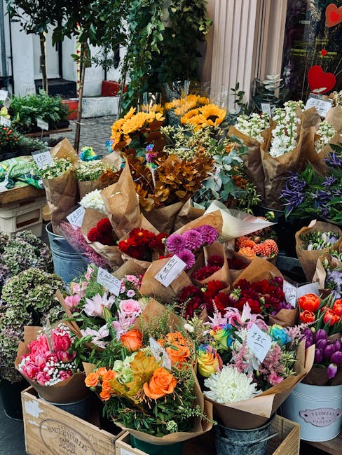 A vibrant display of fresh flower bouquets arranged outdoors in containers and paper wrapping, featuring a variety of colors including pink, orange, red, purple, yellow, and white. The bouquets includ