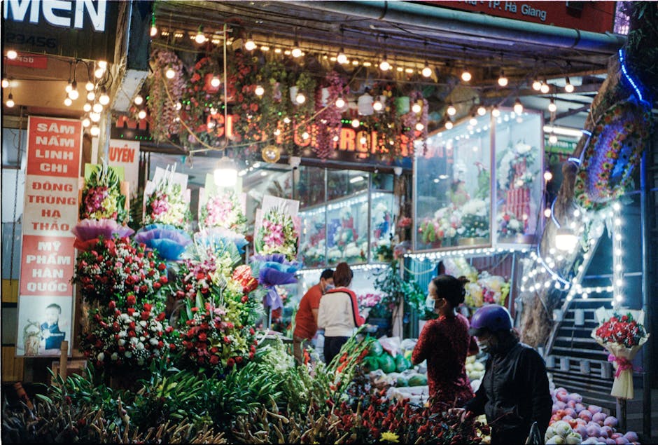 A vibrant flower stall on Homerton High Street at night, illuminated by string lights and decorative lanterns. The display features an assortment of fresh flowers arranged in colorful bouquets, includ
