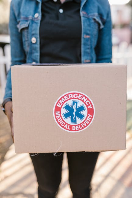 A person holding a rectangular cardboard box with an 'Emergency Medical Delivery' sticker, with only part of their denim jacket and black clothing visible; the background is blurred, suggesting an out